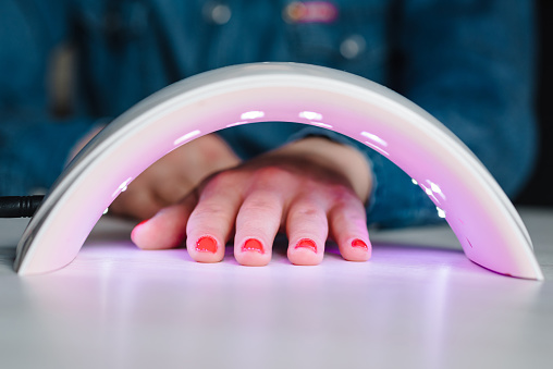 UV lamp with light for drying nails with gel polish. Woman hand inside lamp for nails on table close up. Red nails dried in the lamp. Girl makes a gel manicure at home, doing manicure herself, draws.